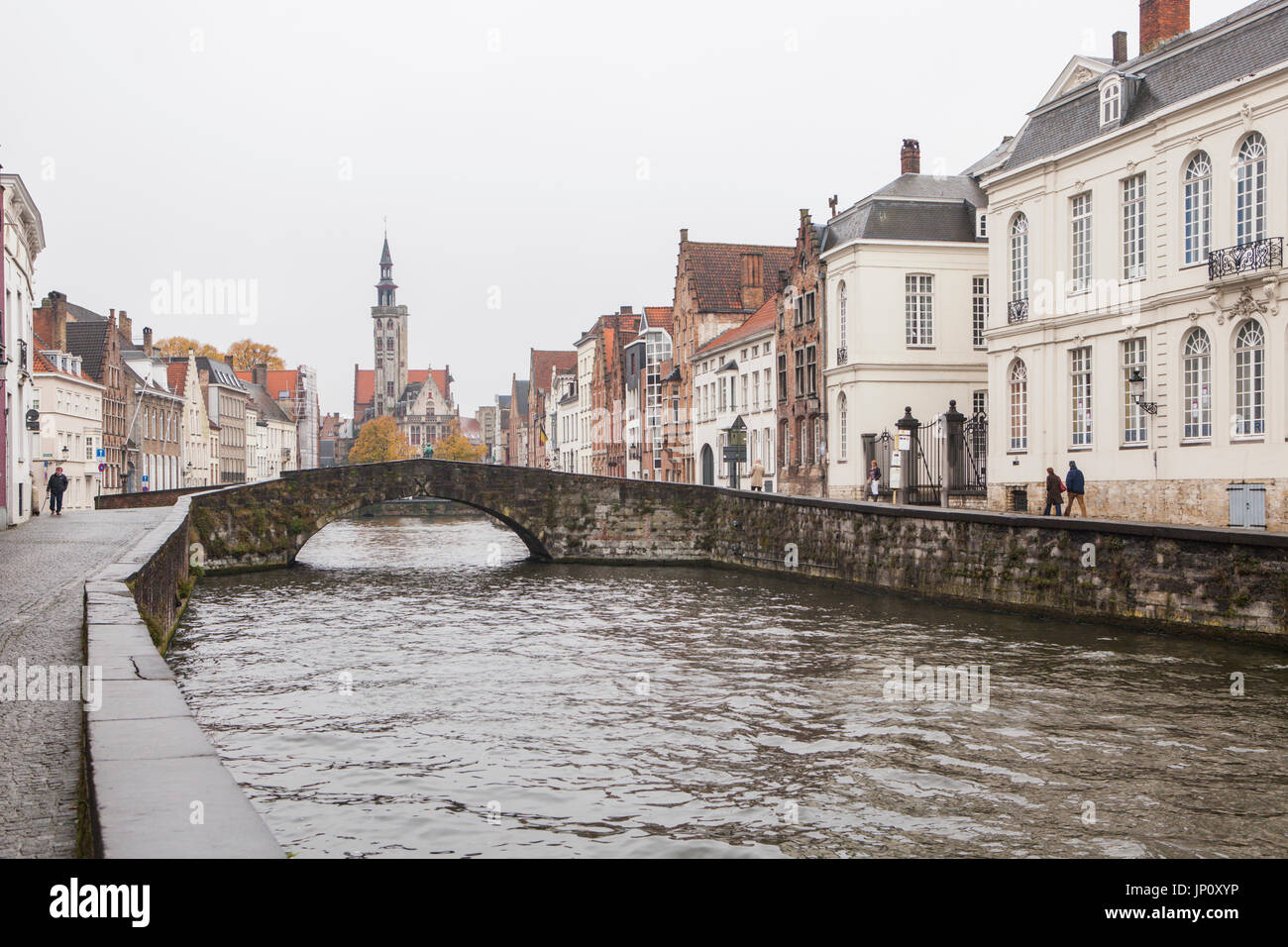 Bruges, Belgium – October 31, 2010: A few pedestrians in the rain on Spinolarei, Koningstraat bridge, the statue of Van Eyck and the Poortersloge beyond. Bruges is sometimes referred to as the Venice of the North. Stock Photo