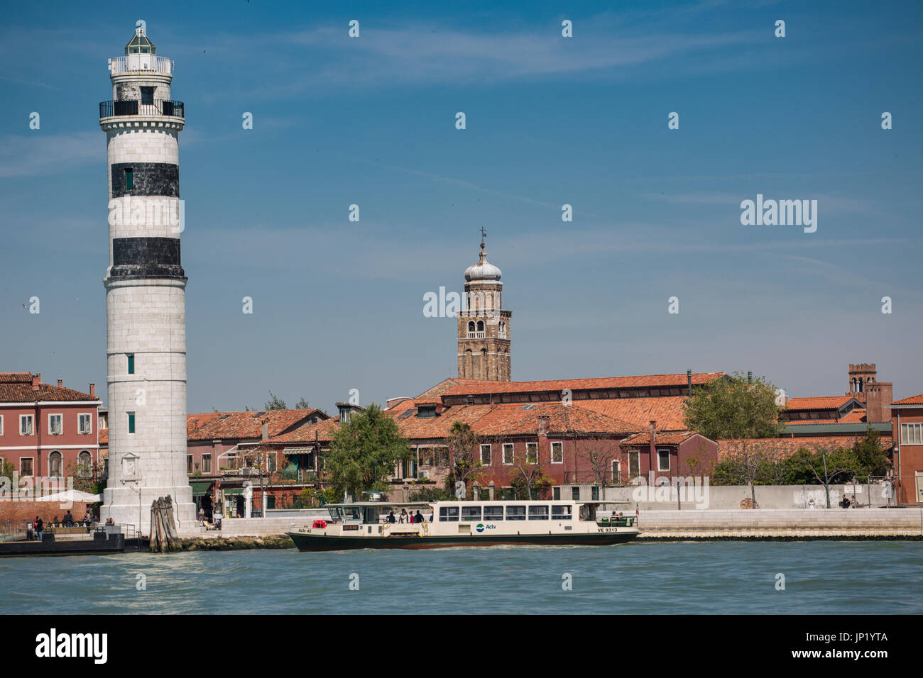 Murano, Venice, Italy - April 28, 2012: The lighthouse (Faro) on Murano in the Venetian Lagoon with the church of Pietro Martire behind. A vaporetto brings and takes off passengers. Stock Photo