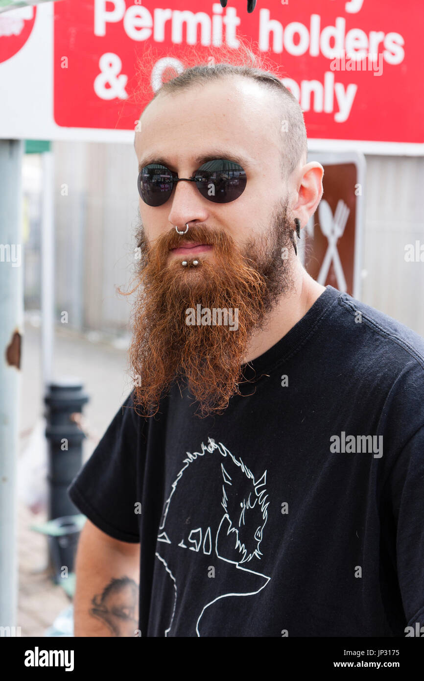 Portrait, face, Caucasian man, 20s, long brown beard, short hair, black round sunglasses. Metal stud piercings chin and nose. Eye-contact. Stock Photo