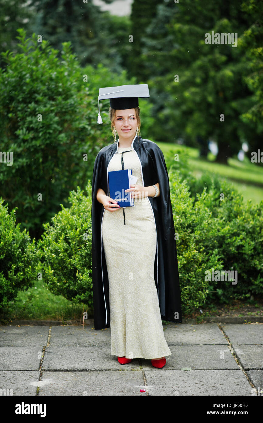 Portrait of a beautiful female graduate in dress and graduation gown ...