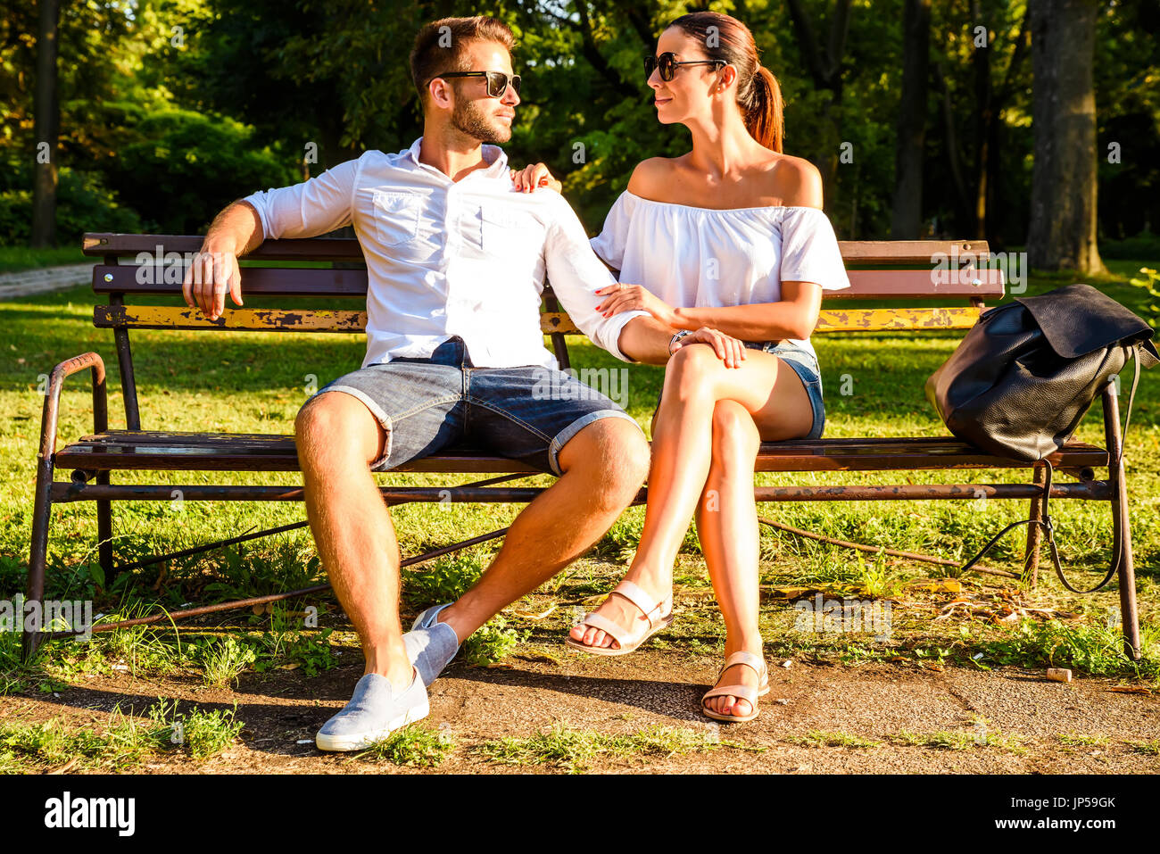 A beautiful young couple sitting on a bench in a park in the sunset ...