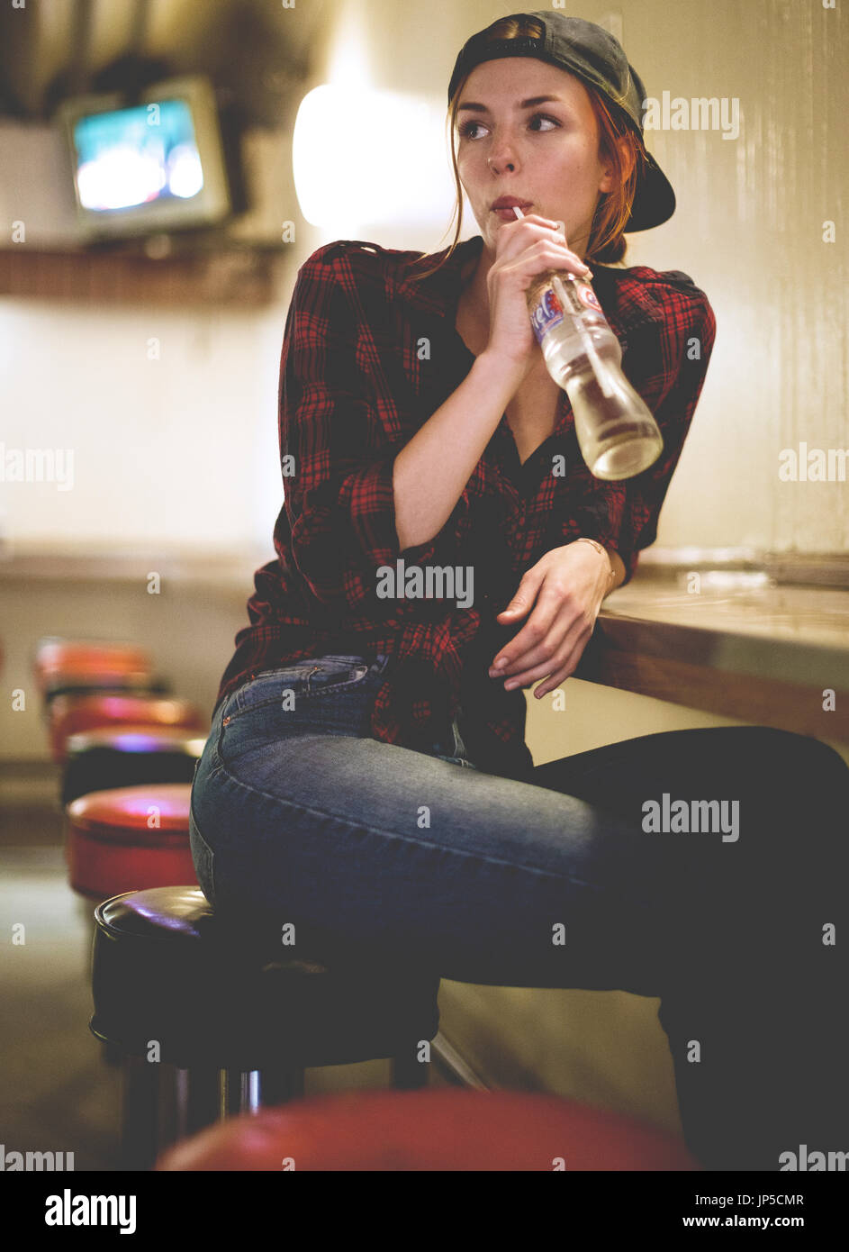 A woman sitting on a stool drinking from a glass bottle in a bar and looking over her shoulder. Stock Photo