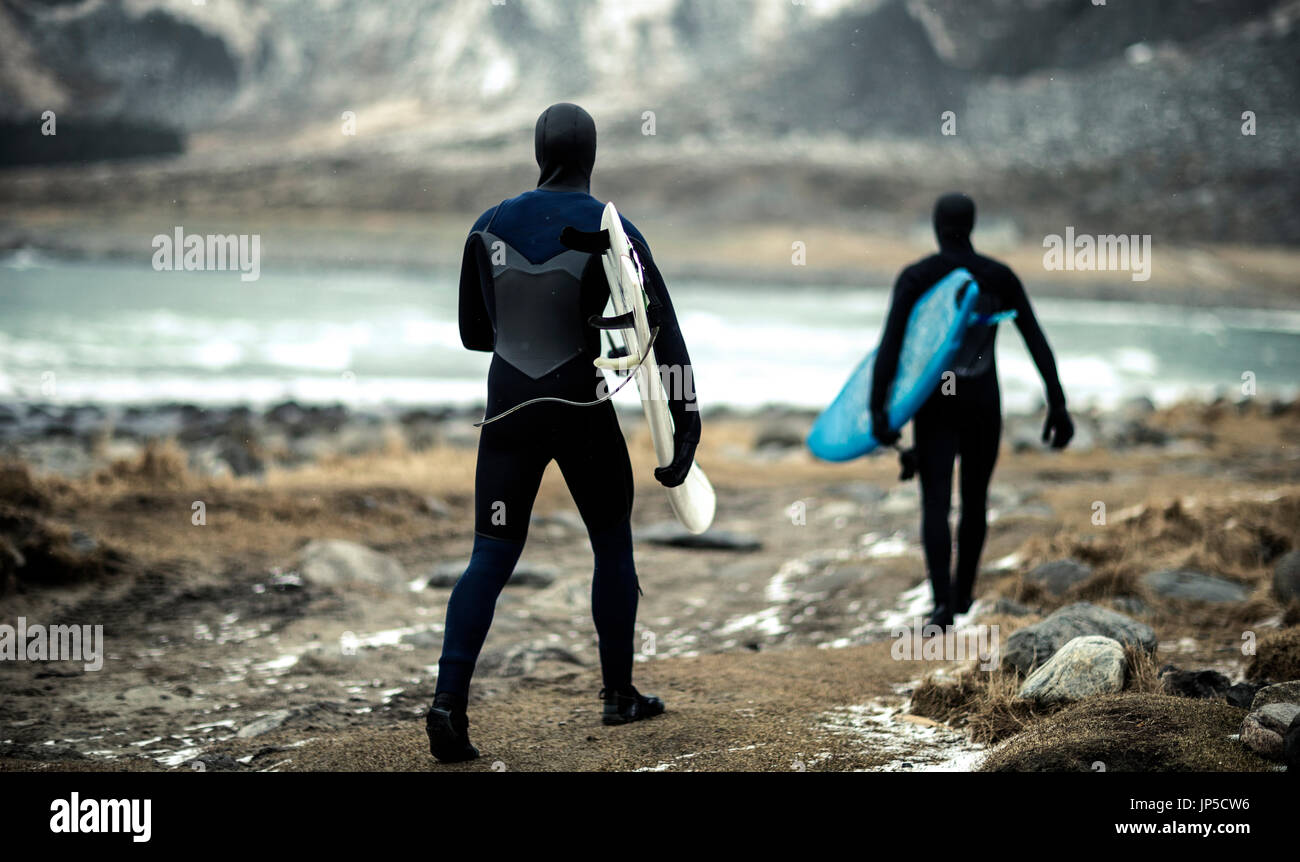 Two surfers wearing wetsuits and carrying surfboards walking towards the ocean with mountains behind. Stock Photo