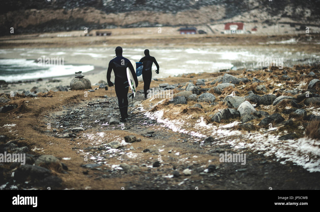 Two surfers wearing wetsuits and carrying surfboards walking along a snowy beach. Stock Photo