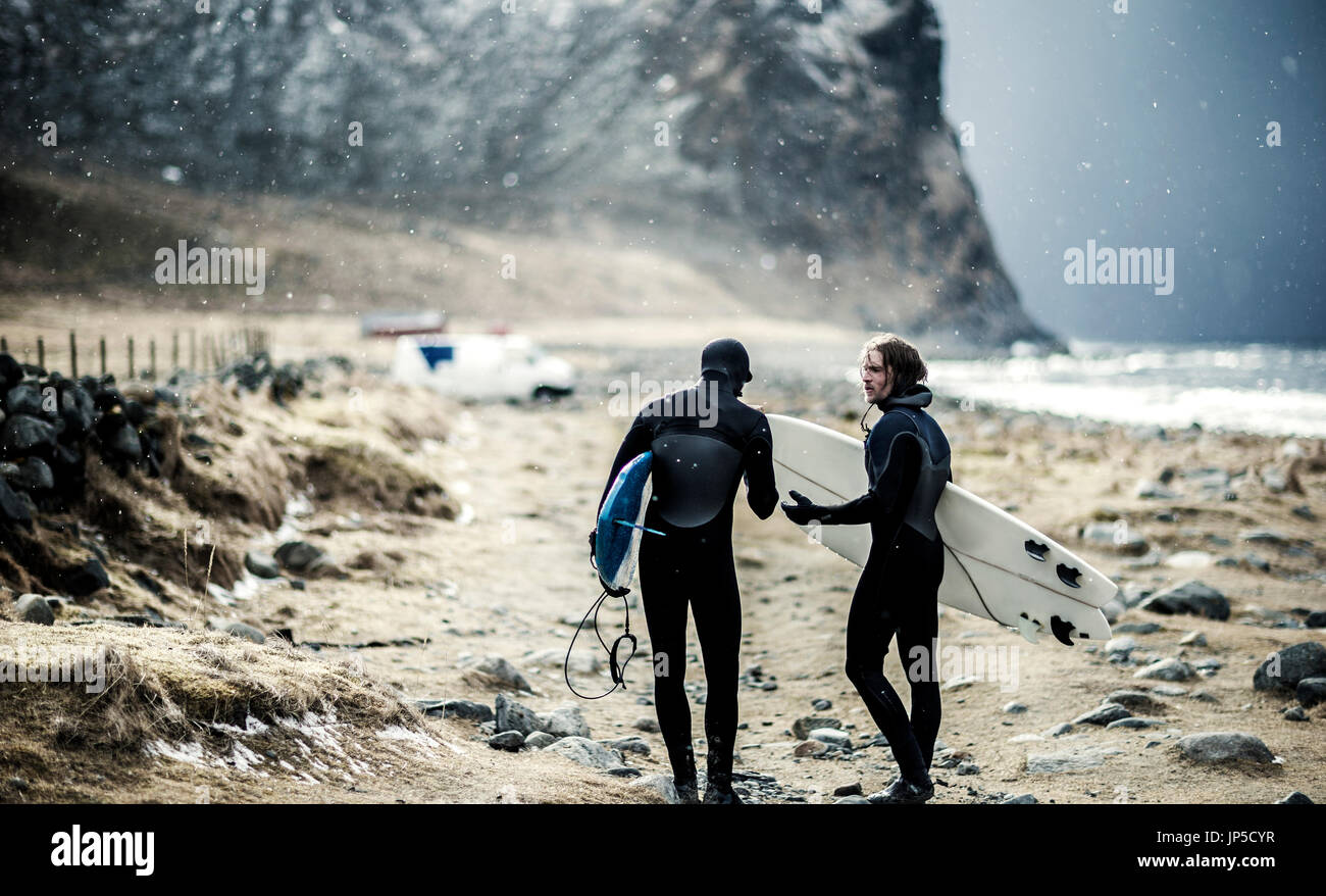 Two surfers wearing wetsuits and carrying surfboards walking towards a van. Stock Photo