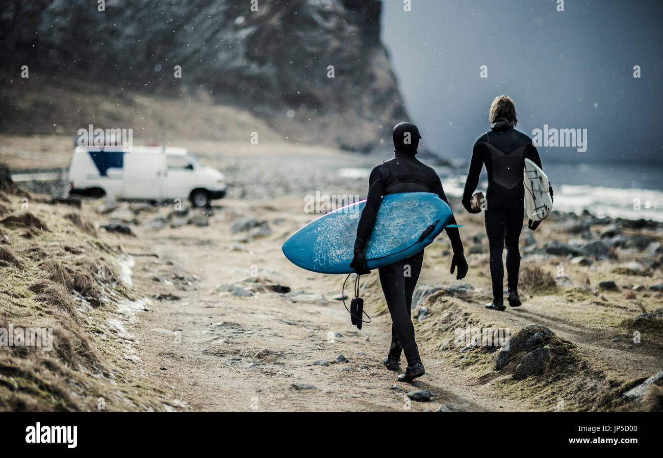 Two surfers wearing wetsuits and carrying surfboards walking towards a van. Stock Photo