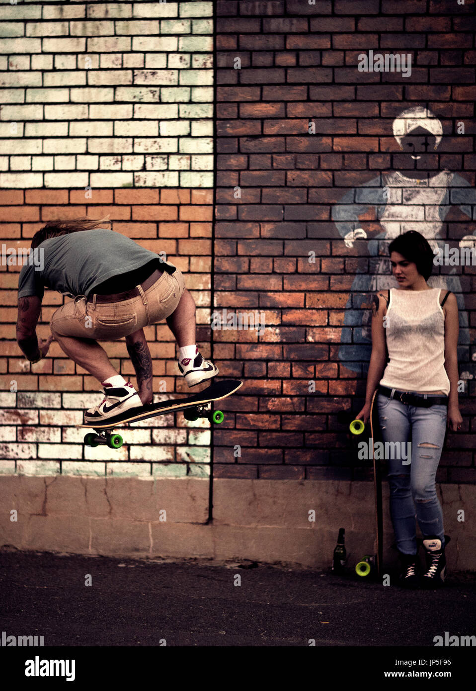 A young man and woman skateboarding in front of a brick wall. Stock Photo