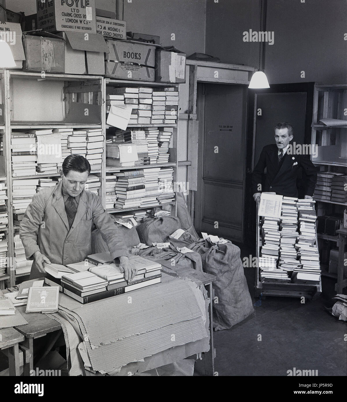 1950s, picture shows a coated employee with tie packing books up ready for despatch in a store room at the famous bookshop of W & G Foyle Ltd, Charing Cross Rd, London, while another suited employee brings more books in on a trolley. Stock Photo