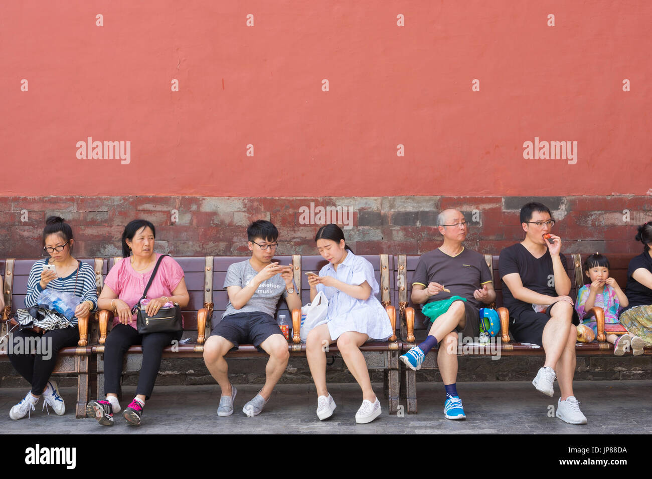 Chinese tourists of different ages sitting on bench at Forbidden City in Beijing - some eating some on their mobile phones and some sitting Stock Photo