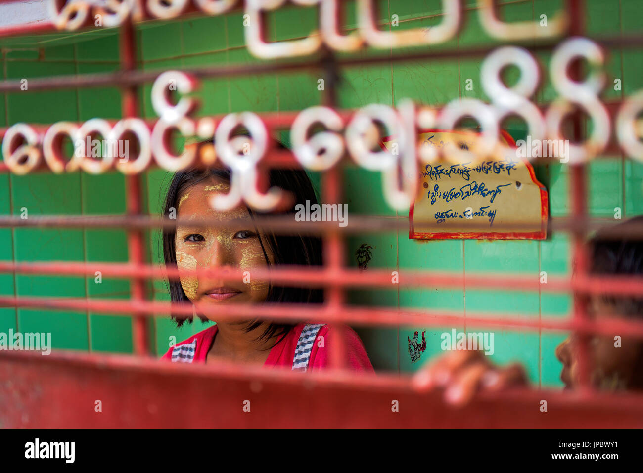 Myngun, Myanmar, South East Asia. Portrait through the gate of a little girl with traditional tanaka makeup. Stock Photo