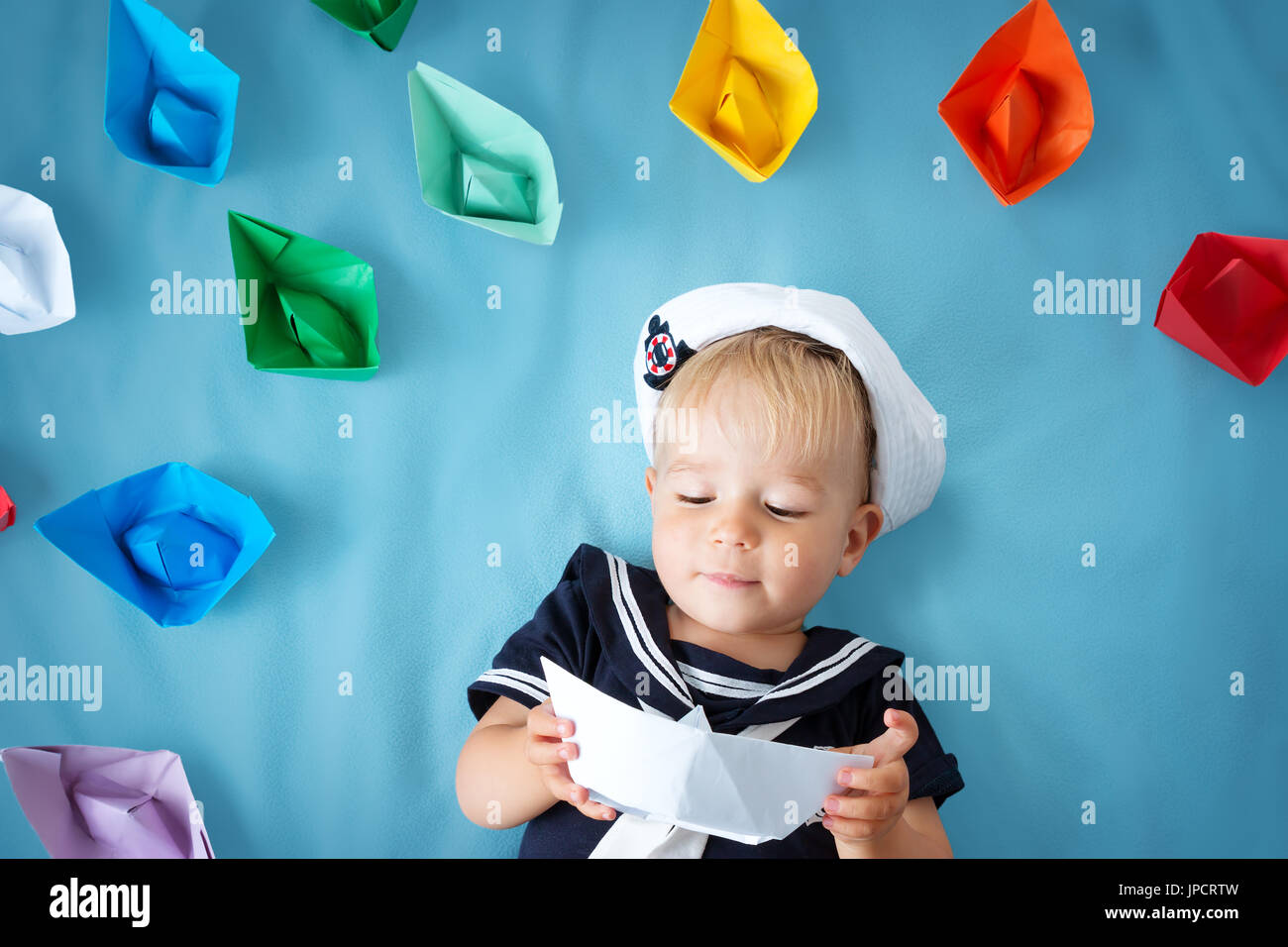 two years old boy playing in sailor hat. Child with paper ships on blue background Stock Photo
