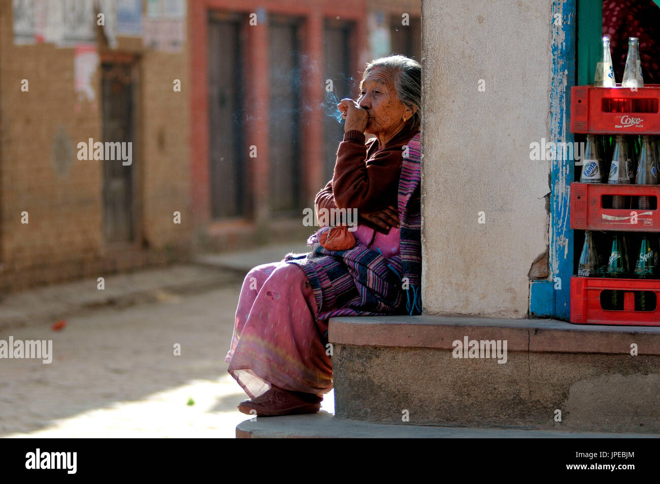 A woman relaxes in the sun, smoking his cigarette and observing the life around her. Panauti, Nepal Stock Photo