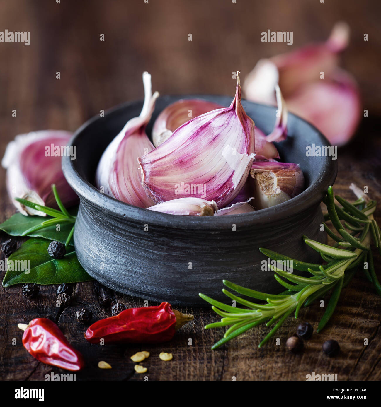 Cloves of purple carlic in a black bowl with rosemary, bay leaf, black pepper and chili pepper on dark rustic wooden background. With plenty copy spac Stock Photo