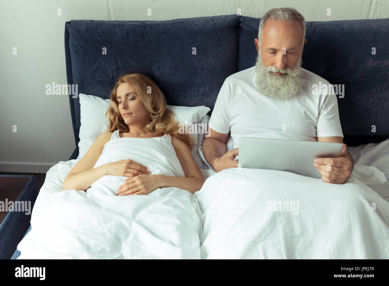 Bearded mature man using laptop in bed while blonde woman sleeping  Stock Photo