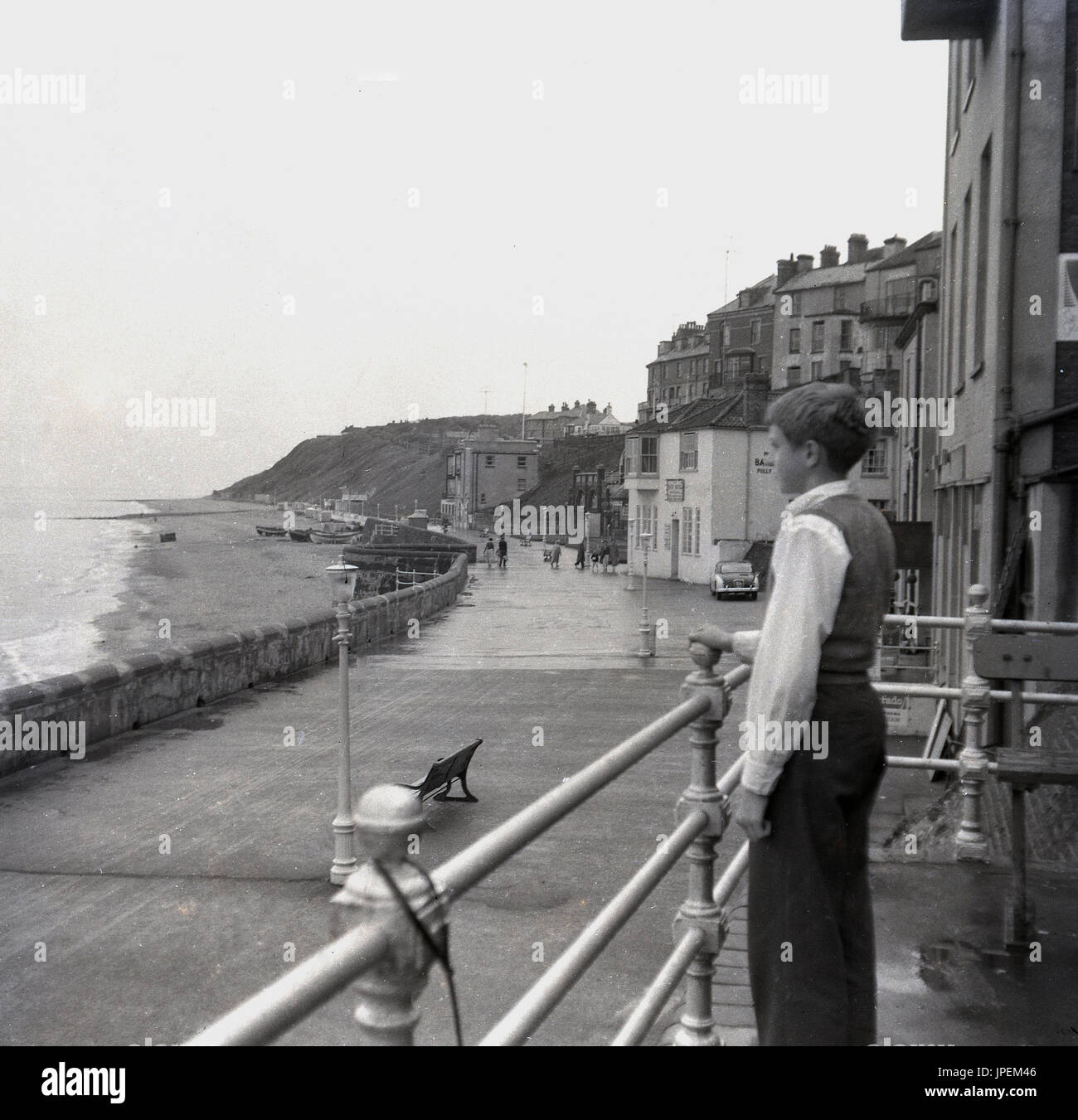 Late 1940s, young boy stands by railings looking over the sea and beach ...