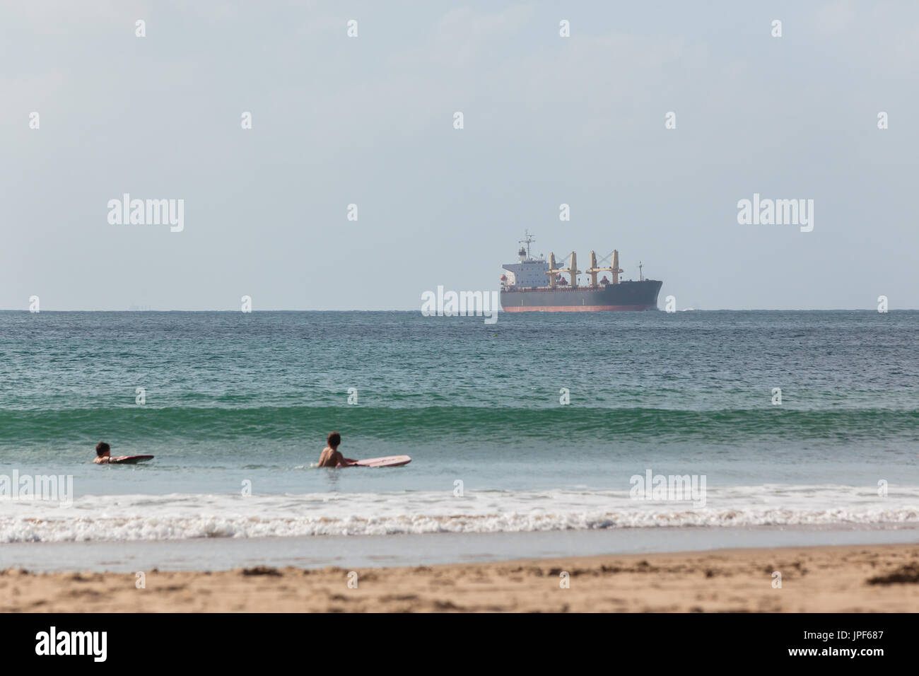Ocean ship enters port closeby beach with young unidentified boys swimming bodyboarding waves. Stock Photo