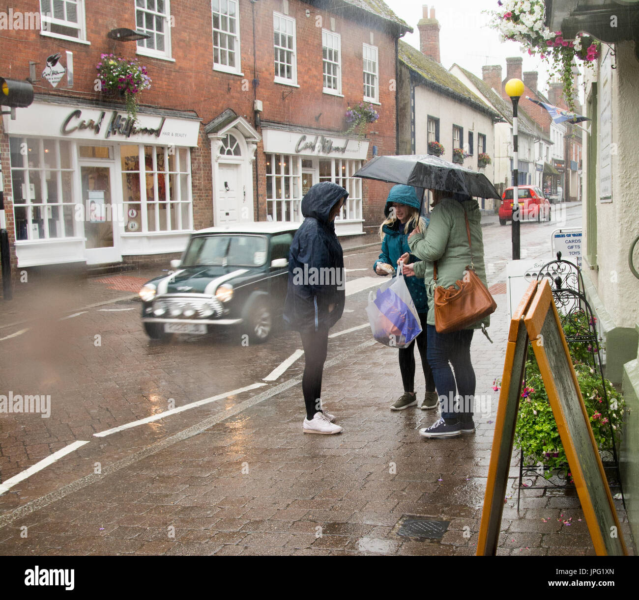 People shopping, talking in English village with umbrellas in rain in summer, August, UK Stock Photo