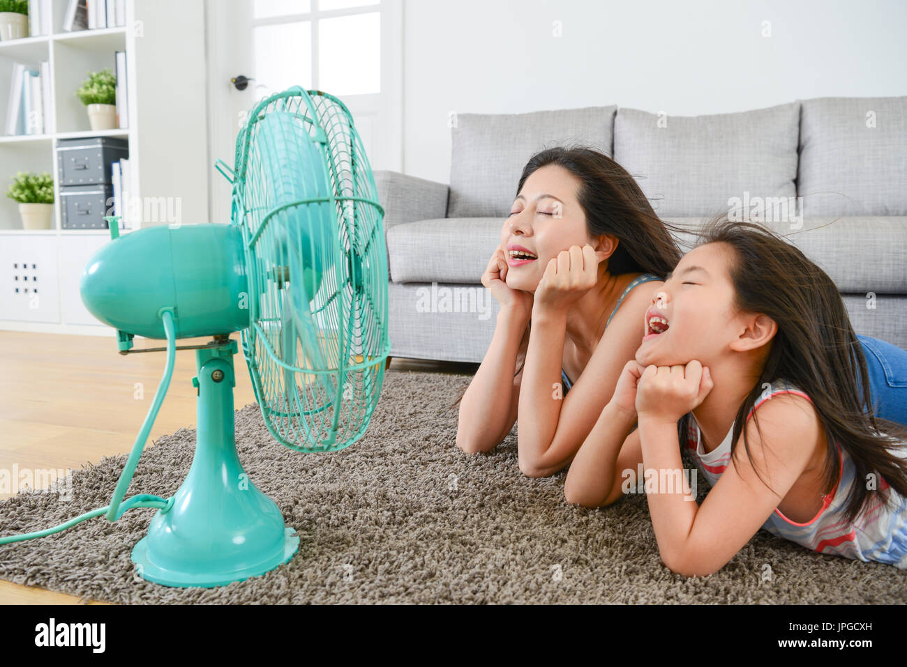 pleasantly mother with young little daughter lying down on living room floor and face to electric fan enjoying blowing cool wind together at summer. Stock Photo