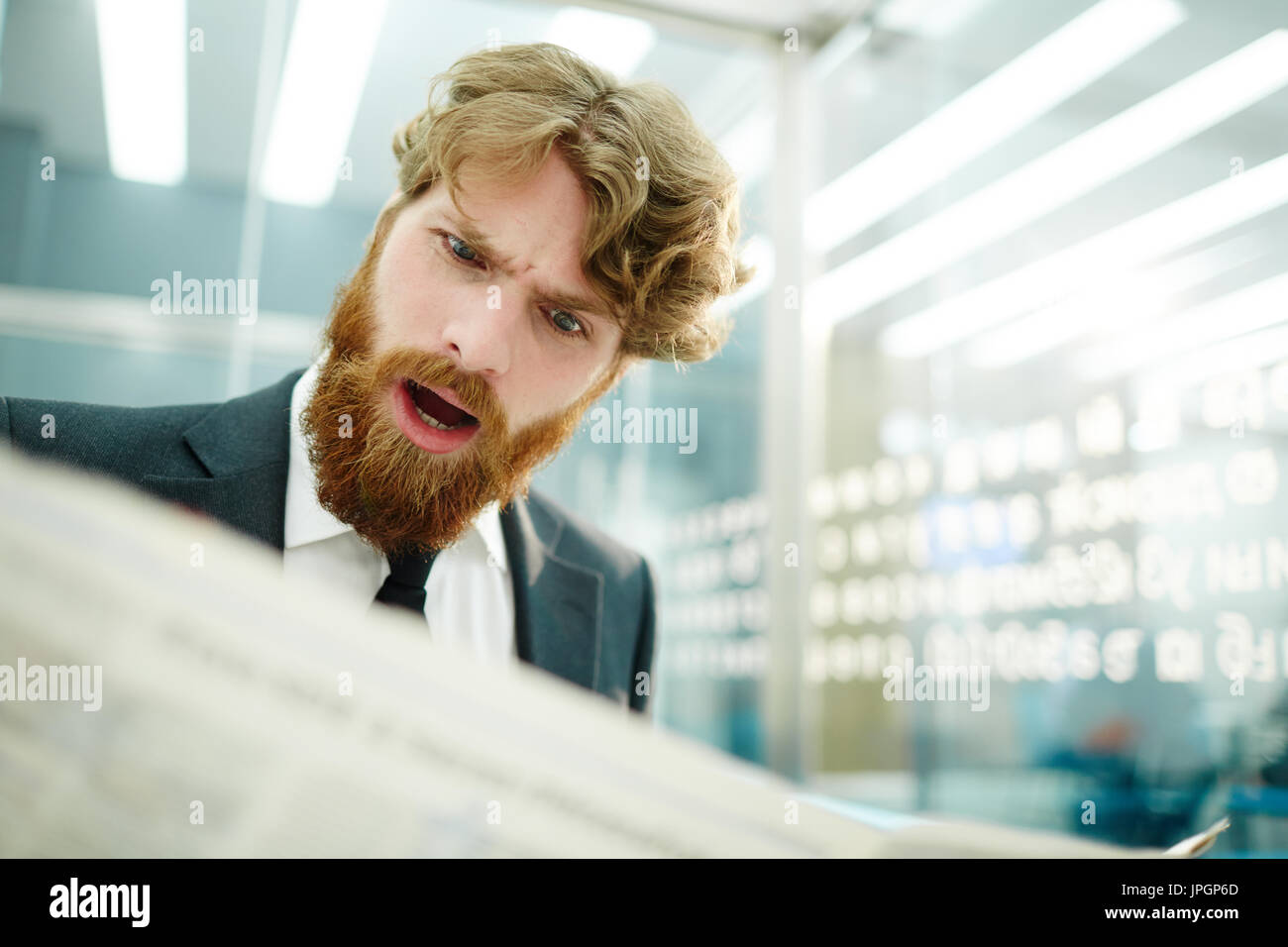 Portrait of shocked bearded businessman reading newspaper in office, frowning with mouth open Stock Photo