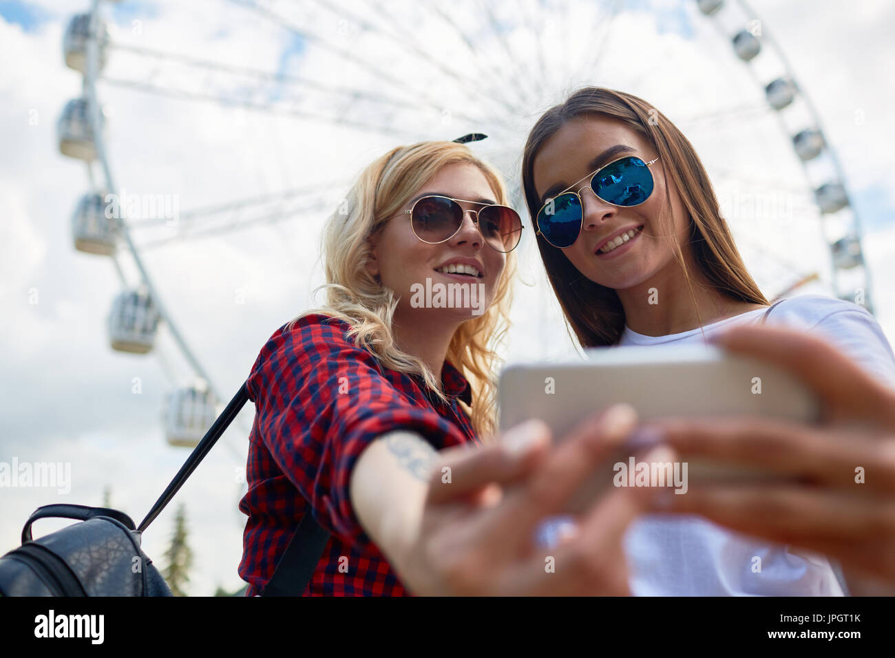 Portrait of two beautiful young girls taking selfie via smartphone camera standing against tall Ferris wheel in amusement park Stock Photo
