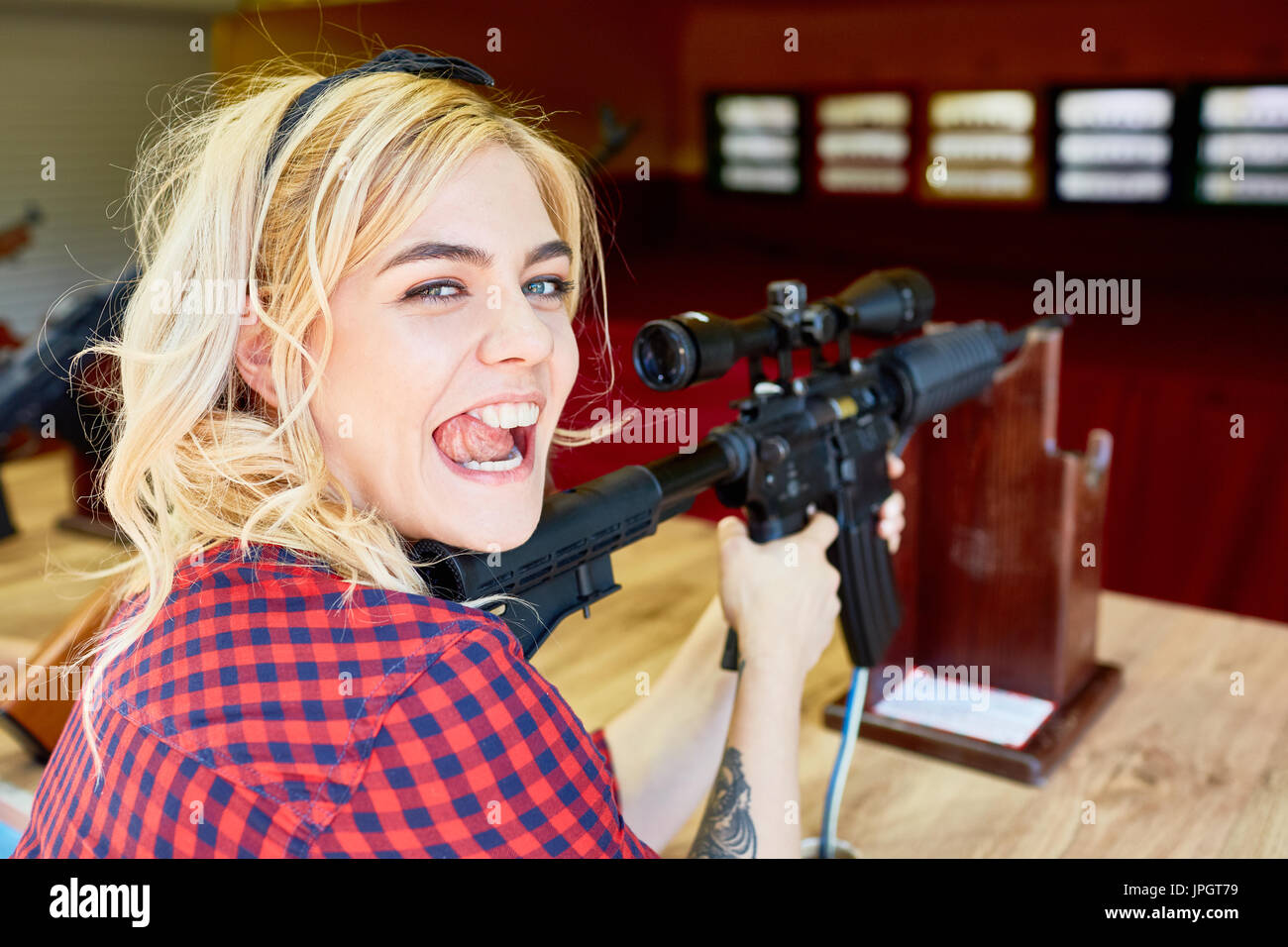 Portrait of cheeky blond girl holding big rifle at shooting stand in amusement park Stock Photo