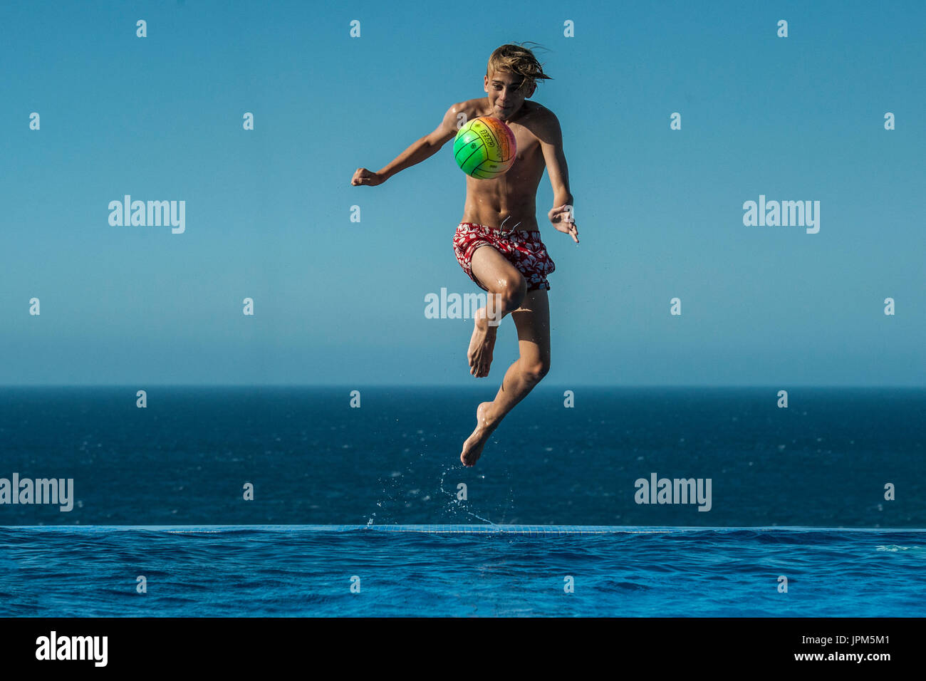 A boy jumps to kick a ball in an infinity pool in the summer, playing football  on holiday Stock Photo