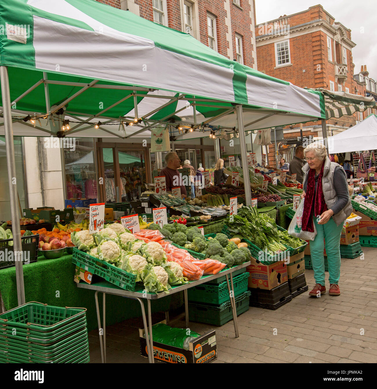 Fruit & vegetable stall with elderly woman looking at colourful fresh produce under green & white awning at street market in Winchester, England Stock Photo
