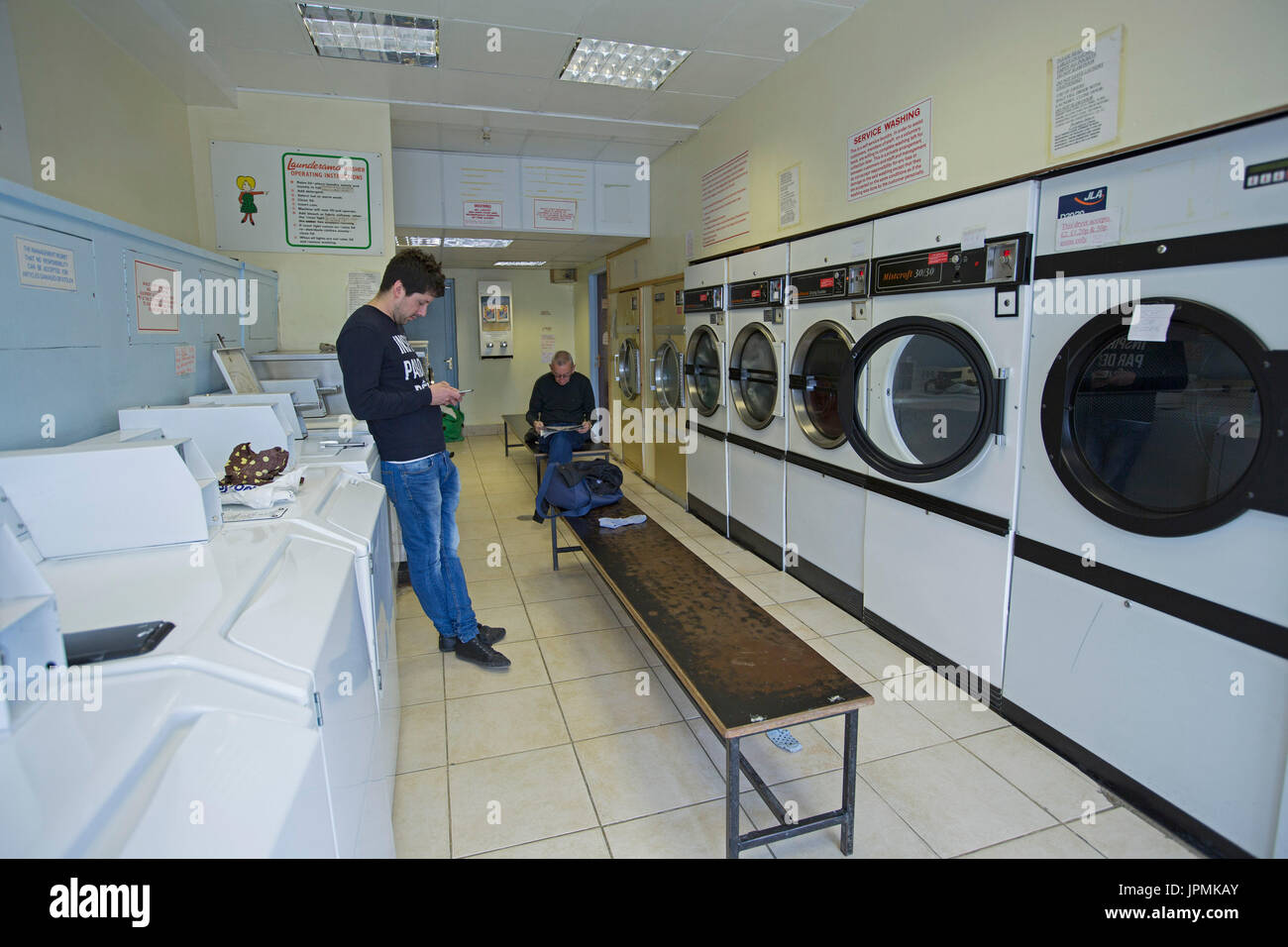 Two men, one reading magazine and one using phone, waiting in laundromat for washing to be completed Stock Photo