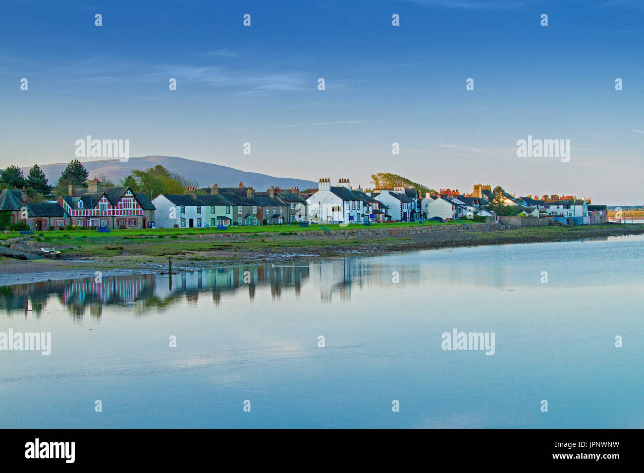 Panoramic view of waterfront houses at coastal village of Ravenglass reflected in mirror surface of blue ocean under blue sky in Cumbria, England Stock Photo