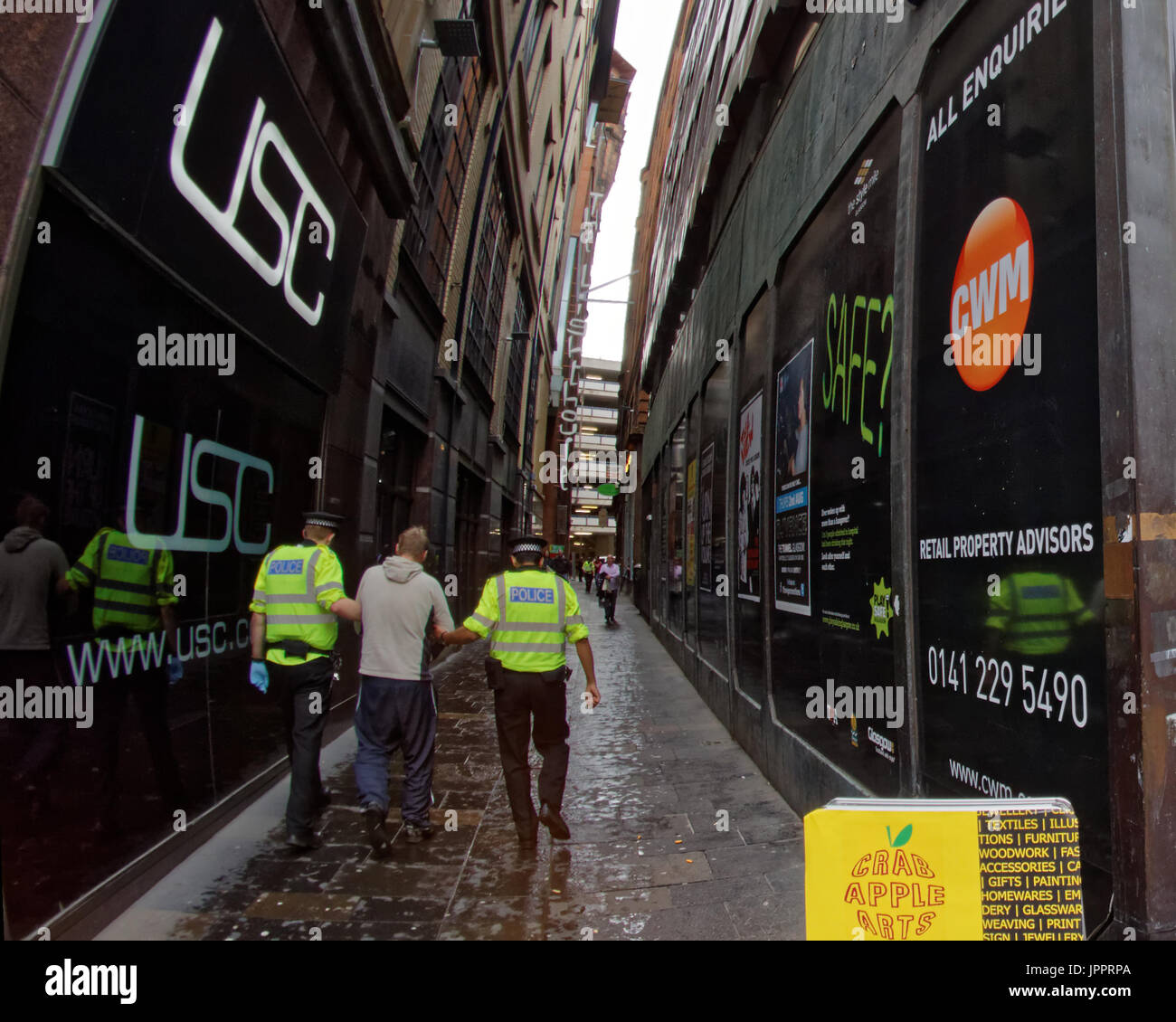 perspective suspect in cuffs being led down an alley by police in high viz jackets surrounded by signs Stock Photo
