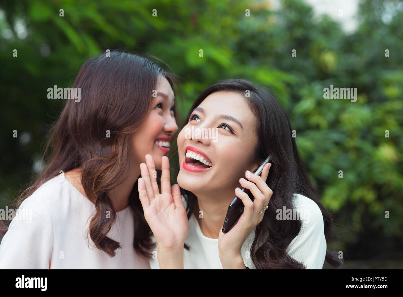 Young woman whispering into cheerful friend's ear while on call Stock Photo