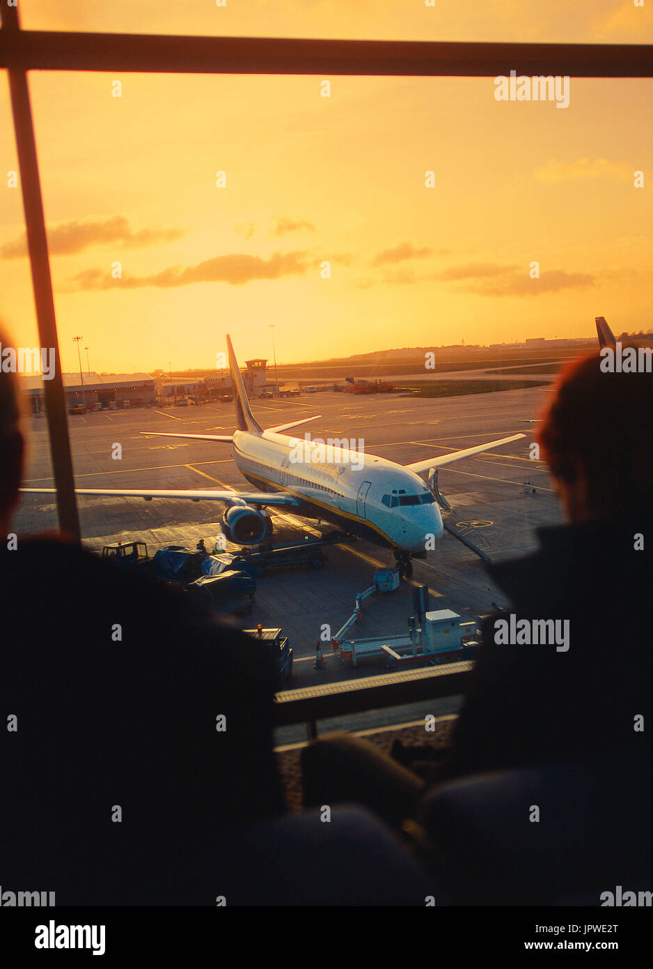 passengers waiting in a full departure-lounge next to windows with a Ryanair Boeing 737-800 parked outside at dusk Stock Photo