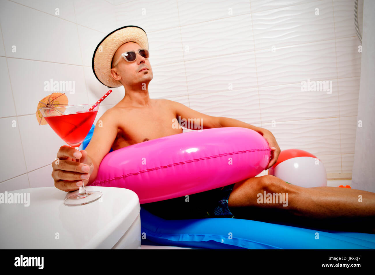 a young caucasian man wearing sunglasses, a straw hat and a pink swim ring pretending he is relaxing in a blue air mattress placed in the shower of a  Stock Photo