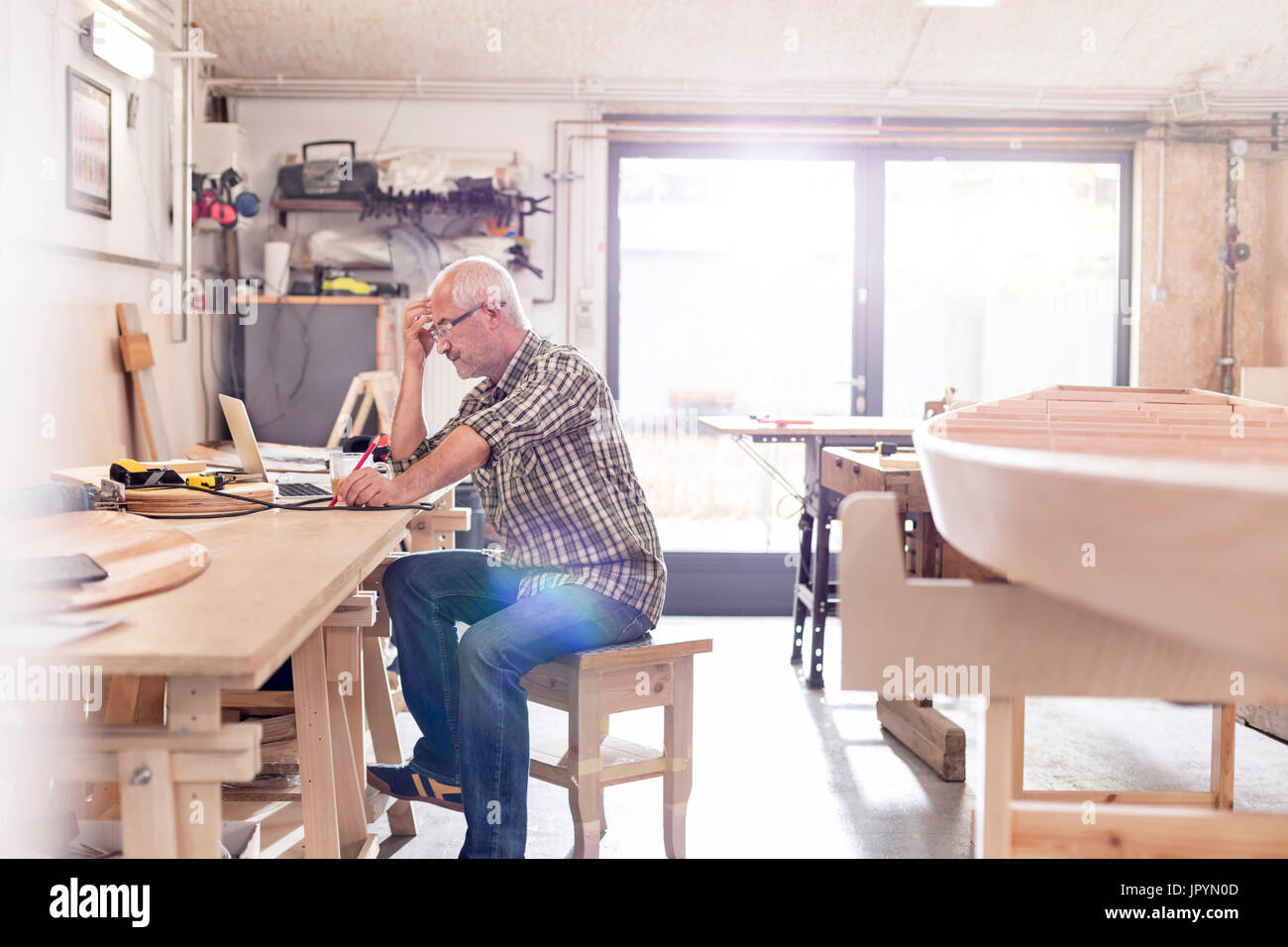 Male carpenter working at laptop on workbench near wood boat in workshop Stock Photo