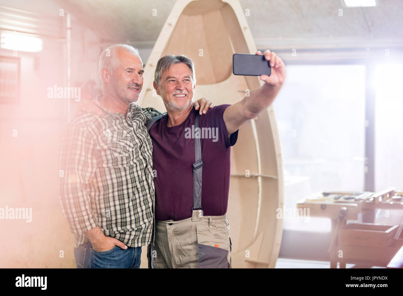 Proud, smiling male carpenters with camera phone taking selfie next too wood boat in workshop Stock Photo