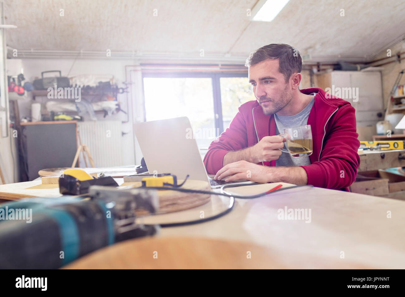 Male carpenter drinking tea and working at laptop on workbench in workshop Stock Photo