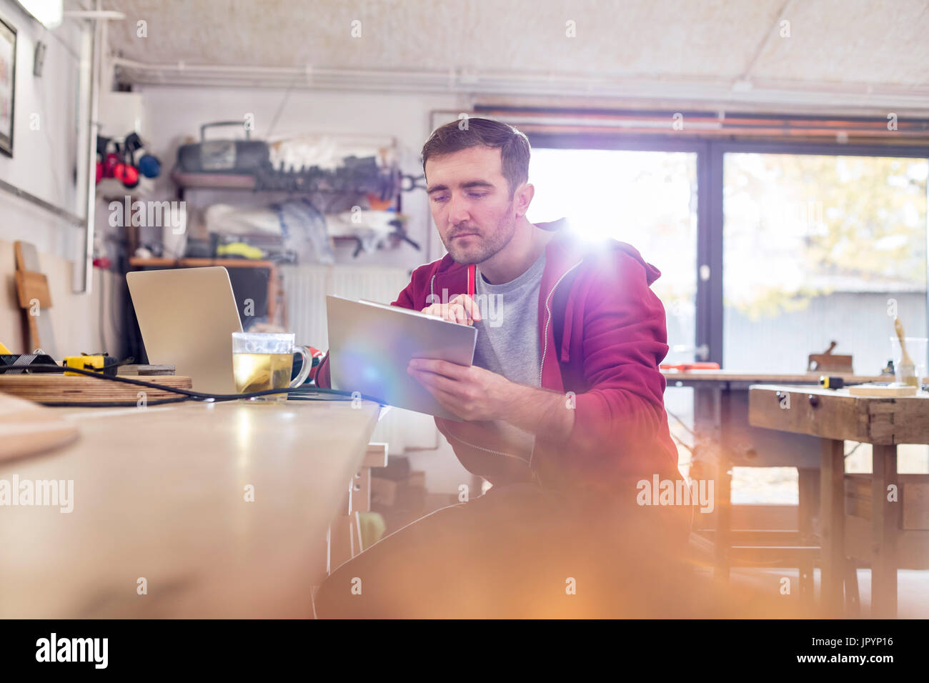 Male carpenter using digital tablet, drinking tea at workbench in workshop Stock Photo