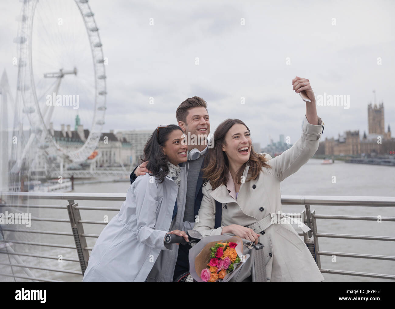 Smiling, happy friends taking selfie with selfie stick on bridge near Millennium Wheel, London, UK Stock Photo