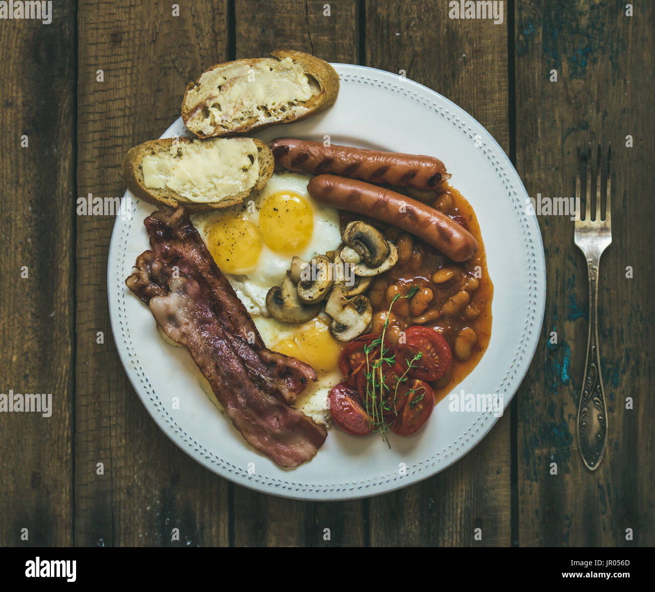 Flat-lay of traditional English breakfast with fried eggs, sausages, mushrooms, sauted tomatoes, bacon, beans and butter toasts on rustic wooden backg Stock Photo