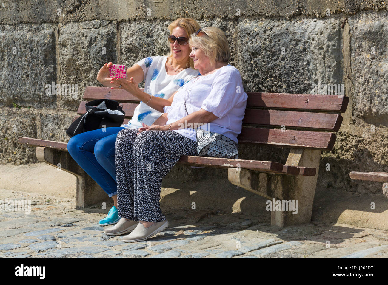Two women sitting on bench taking selfie at Lyme Regis, Dorset UK in July Stock Photo