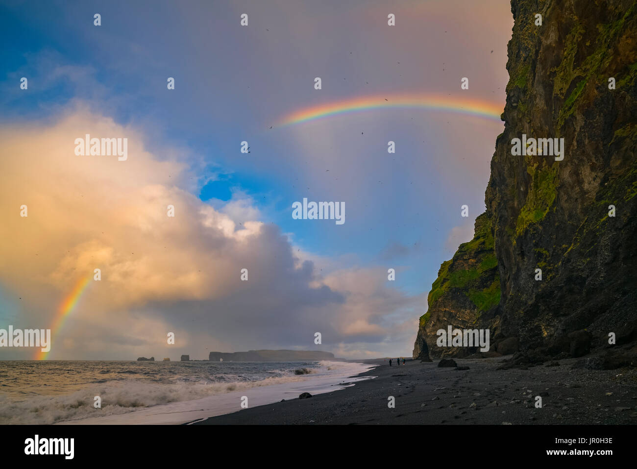 The Sea Stacks Known As Reynisdrangar With Waves At Sunrise, And Thousands Of Birds Flying In The Air With A Rainbow, South Coast; Iceland Stock Photo