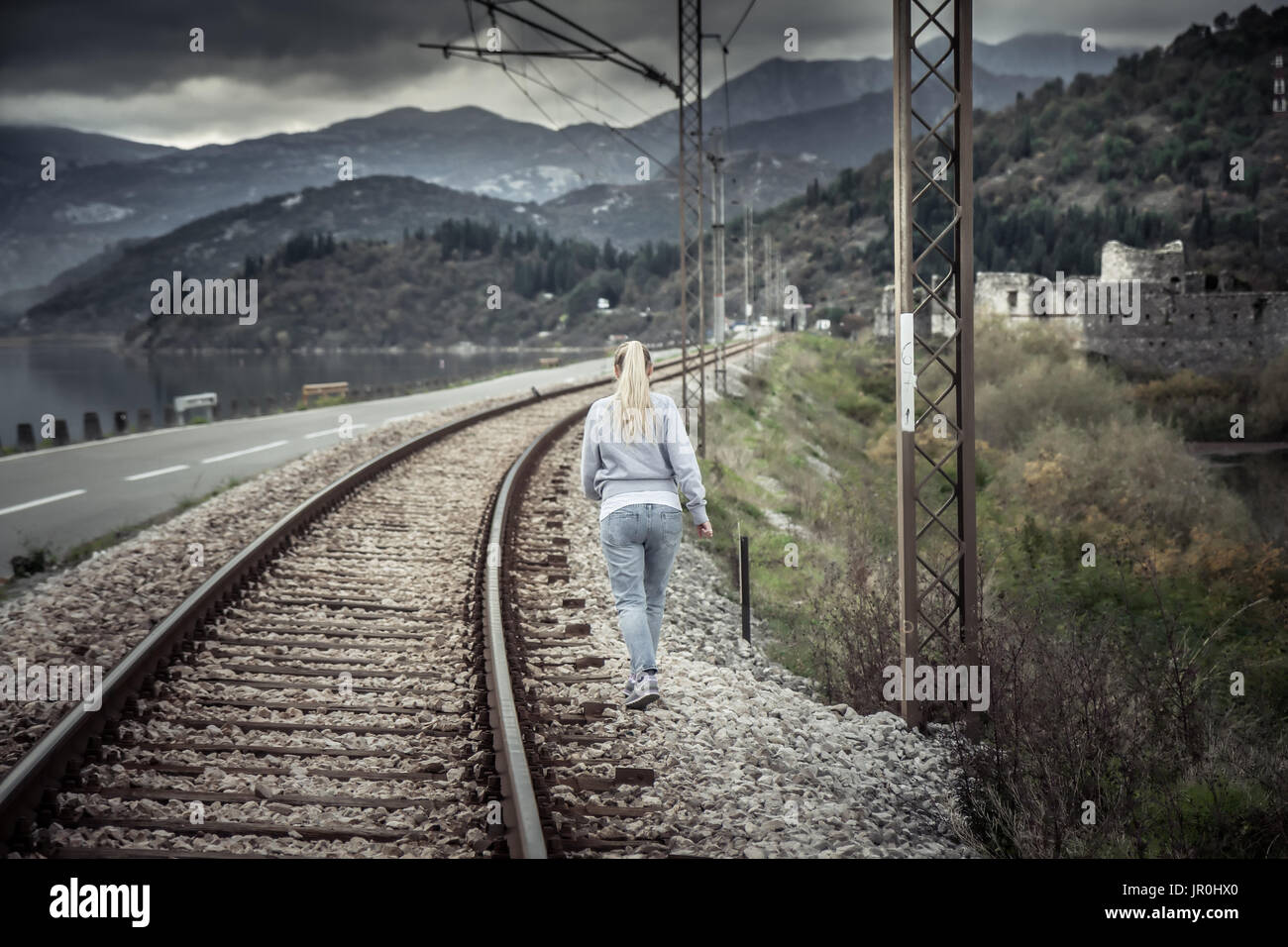 Pensive young woman going away into the distance across railway path in overcast day with dramatic sky  symbolizing parting and despair in relationshi Stock Photo