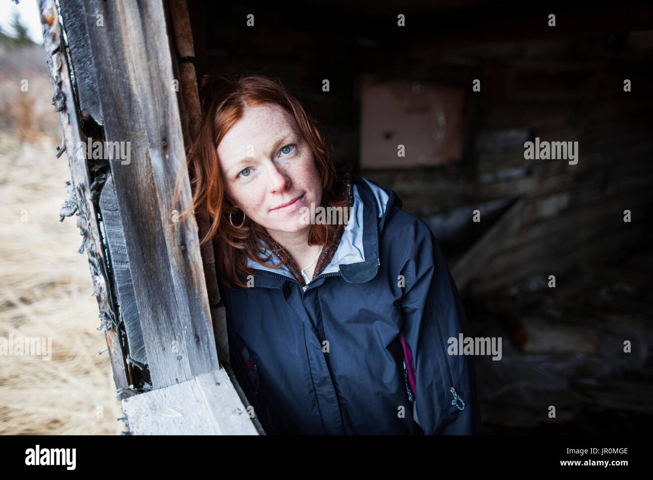 Portrait Of A Woman With Red Hair Leaning Against A Wooden Wall And Looking Up At The Camera; Alaska, United States Of America Stock Photo
