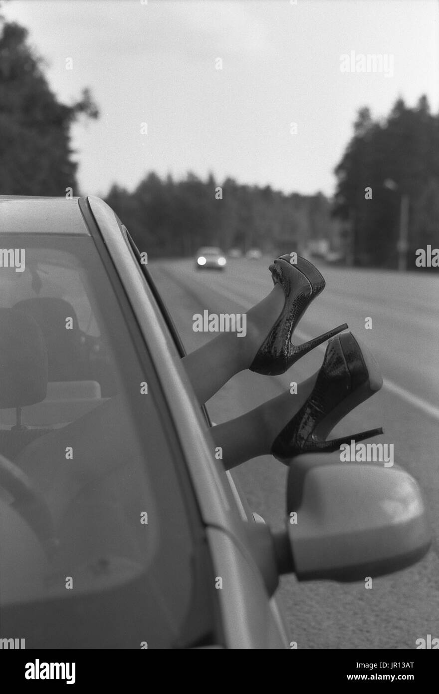Female feet in high-heeled shoes stick out of the car. Black and white photo. Attention! The image contains the graininess of the photographic film! Stock Photo