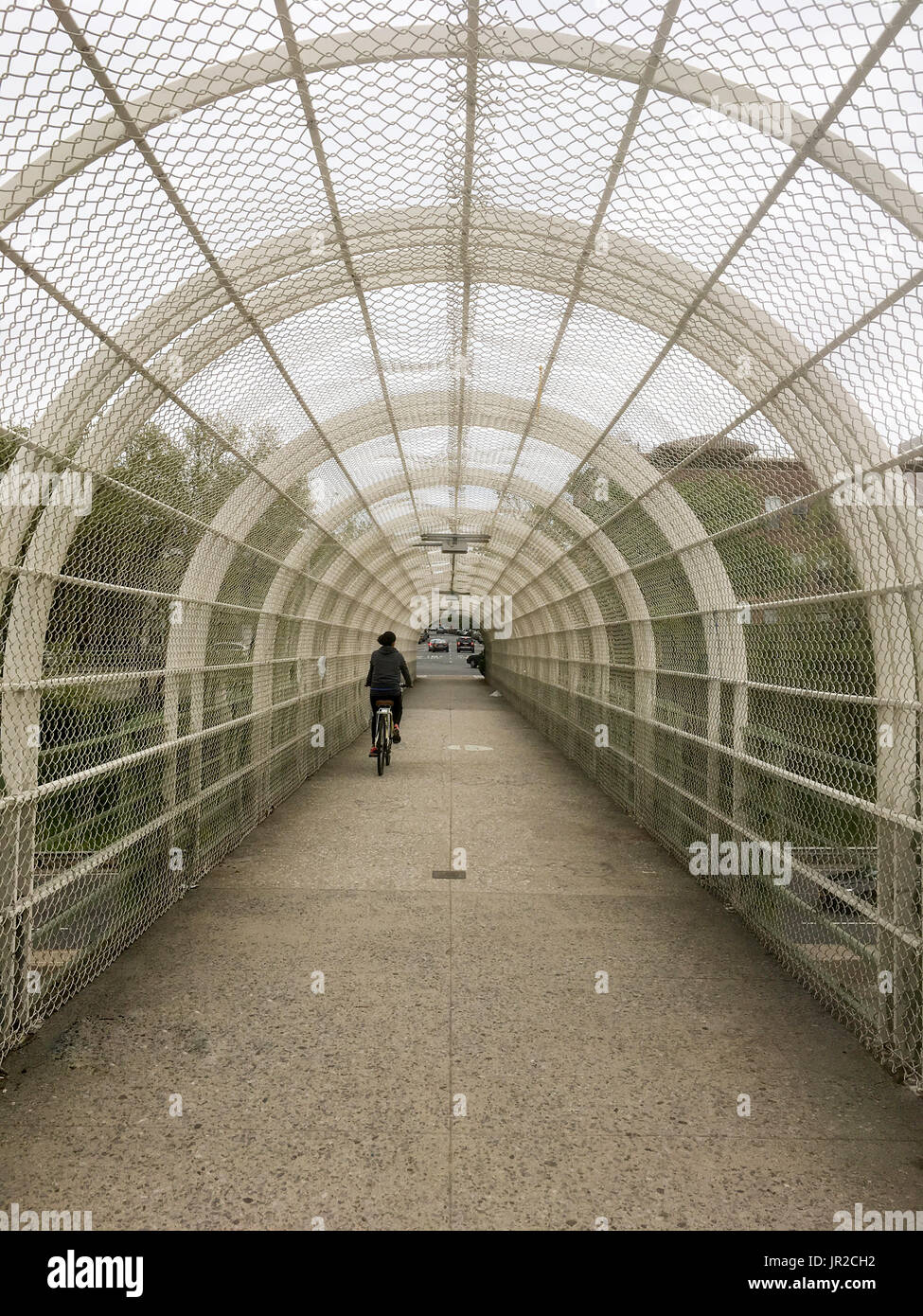One woman riding a bike wearing black on a covered overpass going over a parkway in Brooklyn New York. Chicken wire covered overpass, partial shelter  Stock Photo