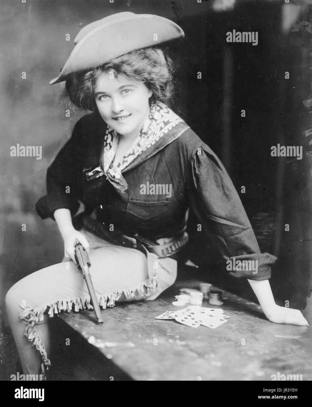 Woman posing in cowgirl outfit, holding revolver, seated on table, with playing cards. The history of women in the west, and women who worked on cattle ranches in particular, is not as well documented as that of men. It wasn't until the advent of Wild West Shows that 'cowgirls' came into their own. These adult women were skilled performers, demonstrating riding, expert marksmanship, and trick roping that entertained audiences around the world. Women such as Annie Oakley became household names. By 1900, skirts split for riding astride became popular, and allowed women to compete with the men wi Stock Photo
