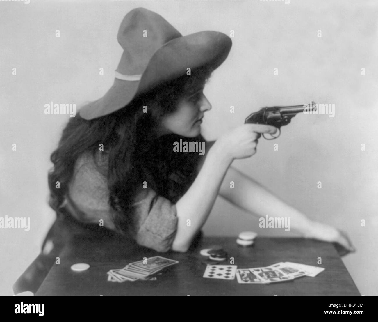 Woman in cowgirl clothing, seated at table with deck of cards and chips, pointing pistol, with arm on table. The history of women in the west, and women who worked on cattle ranches in particular, is not as well documented as that of men. It wasn't until the advent of Wild West Shows that 'cowgirls' came into their own. These adult women were skilled performers, demonstrating riding, expert marksmanship, and trick roping that entertained audiences around the world. Women such as Annie Oakley became household names. By 1900, skirts split for riding astride became popular, and allowed women to c Stock Photo