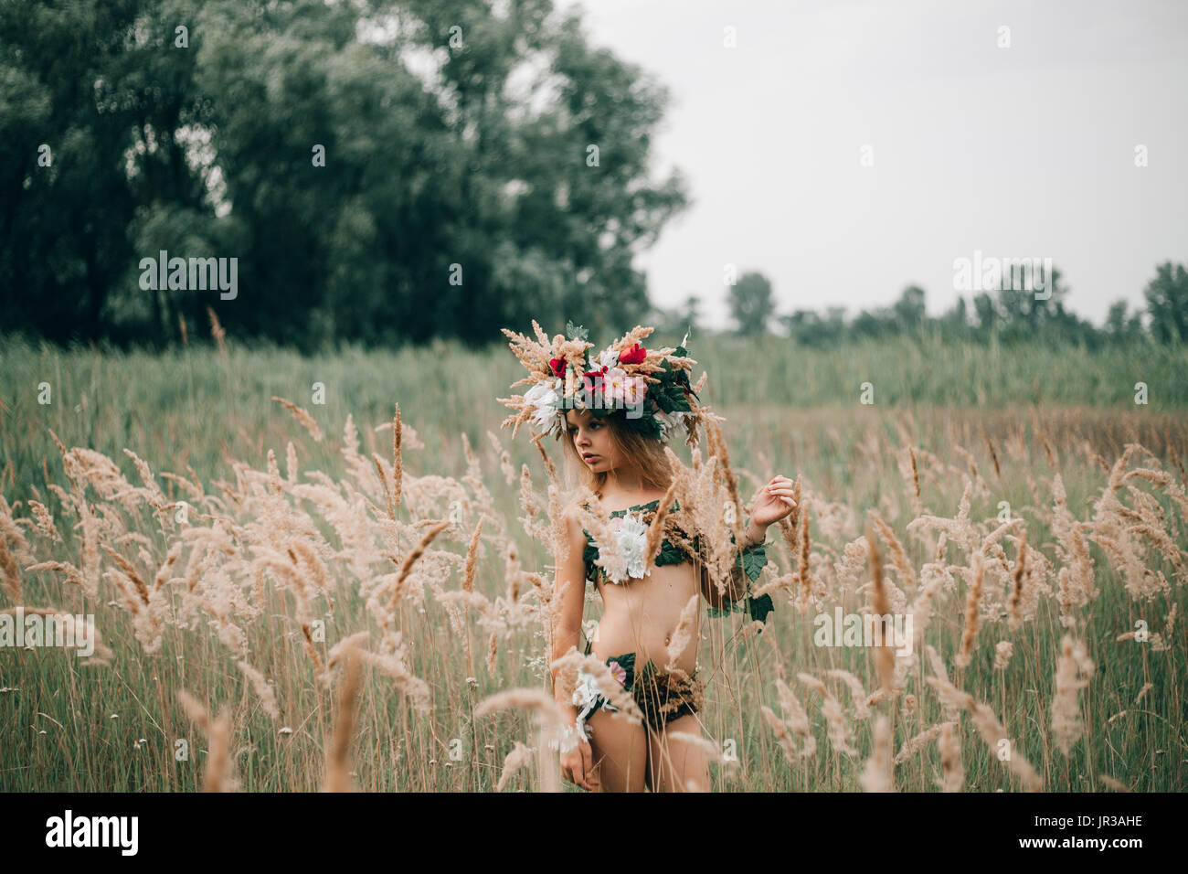 Beautiful little girl in image of nymph with floral head wreath stands among grasses on meadow. Stock Photo