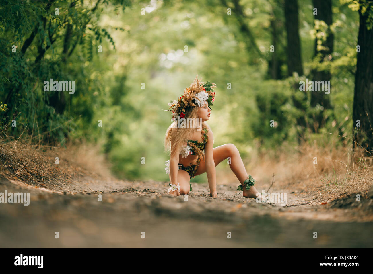 Beautiful little girl in image of nymph dryad with floral head wreath sits in forest road. Stock Photo