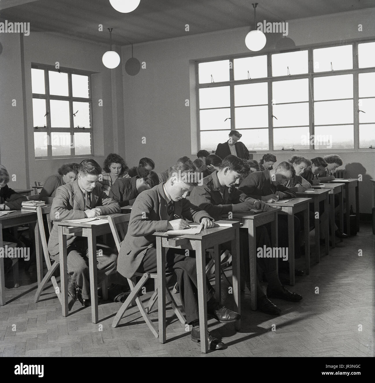 1950s, England, young adult students sitting at small individual wooden desks in a classroon at a technical college. They are writing on paper with pens, while a female teacher monitors the work, England, UK. The young males are all wearing jacket & ties, a formal standard of dress common to this post-war era. Stock Photo